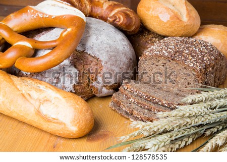 Different types of bread and bakery products such as a loaf of bread, pretzels, buns and slices of whole grain bread lying on a table with a bunch of wheat