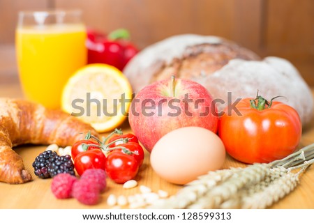 Different types of food such as bread, a tomato, apple, pine seeds, raspberry and a croissant as well a glass of orange juice