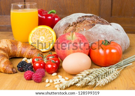 Different types of food such as bread, a tomato, apple, pine seeds, raspberry and a croissant as well a glass of orange juice