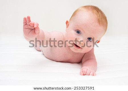 Six month old white baby boy, with blue eyes, lying on his belly on a white matras and lifting his arm.