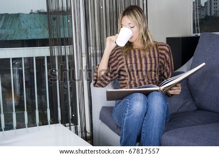 happy blond woman on her couch reading a book and taking a sip from her coffee