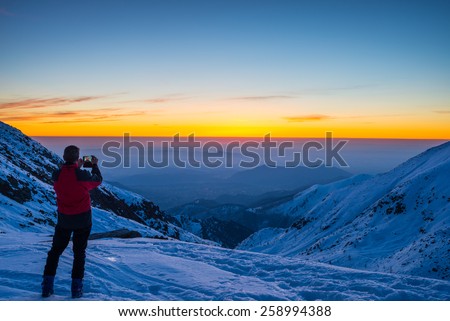 Alpinist with phone in hand taking selfie on the mountain summit with majestic panoramic view of the Alps during dusk time. Concept of sharing life moments using new technology and wireless connection