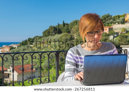 Mature lady with glasses and casual clothings working at laptop outdoors on terrace. Beautiful background of green hills and blue sky in a bright sunny morning. Natural daylight, real people.