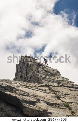 Hiker climbing on steep rocky slope towards the mountain summit without protection. Dramatic cloudy sky. Concept of reaching the goal and conquering the success.