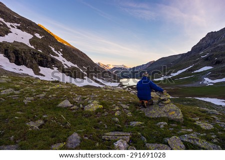 One person sitting on rocky terrain and watching a colorful sunrise high up in the Alps. Wide angle view from above with glowing mountain peaks in the background. Summer adventure and exploration.