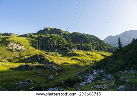 High voltage power line crossing idyllic alpine environment hit by the first sunlight of the day. Summer adventures in the Italian Alps. Wide angle view.