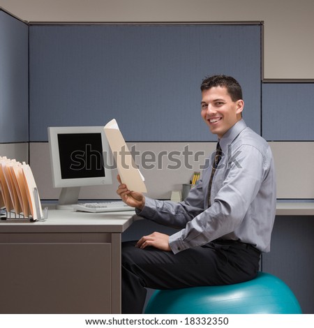 Smiling businessman sitting on exercise ball at desk in cubicle
