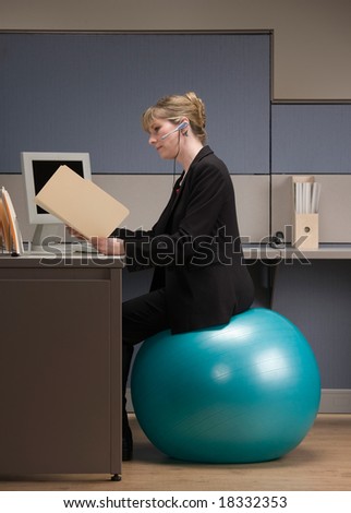 Businesswoman in headset looking at file folder while sitting on exercise ball at desk in cubicle