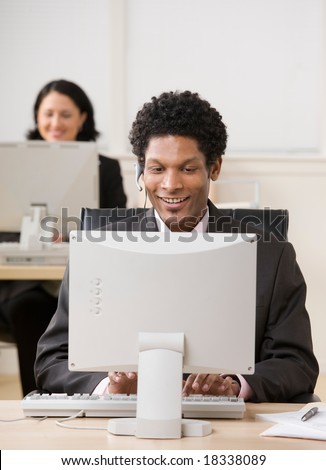 Happy businessman in headset working on computer with co-worker in background