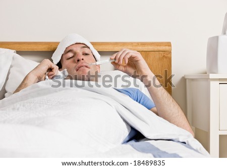 Sick man with fever laying in bed taking temperature with thermometer