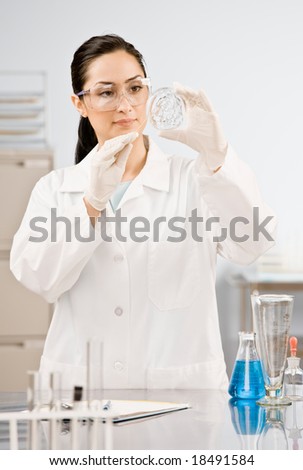 Research scientist in lab coat, safety goggles and rubber gloves testing specimen in petri dish in laboratory