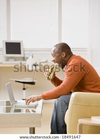 Hungry man multi-tasking by eating a sandwich and typing on laptop in livingroom