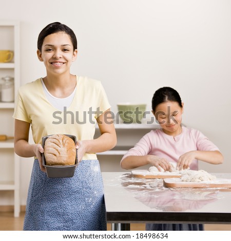 Girl displays baked loaf of bread while sister kneads dough
