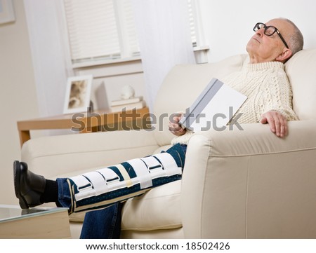Disabled man with leg brace holding book and napping on sofa in livingroom