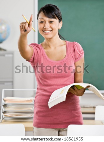 Happy teacher with text book gesturing and asking questions in school classroom