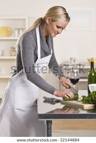 Attractive blond woman slicing cucumber in kitchen while drinking glass of wine looking down.  Vertical