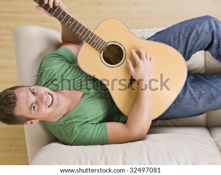 An above view of an attractive young man lying on the couch and playing a guitar.  He is smiling directly at the camera.  Horizontally framed shot.