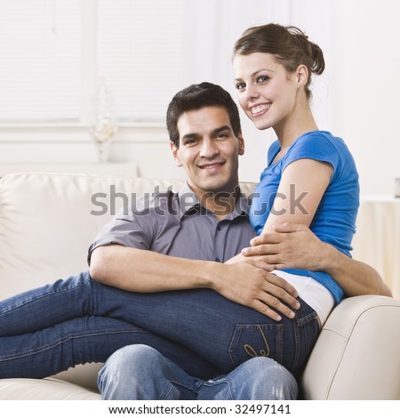 An attractive young couple relaxing together in their home.  The woman is sitting on the man\'s lap and they are smiling at the camera happily. Square composition.