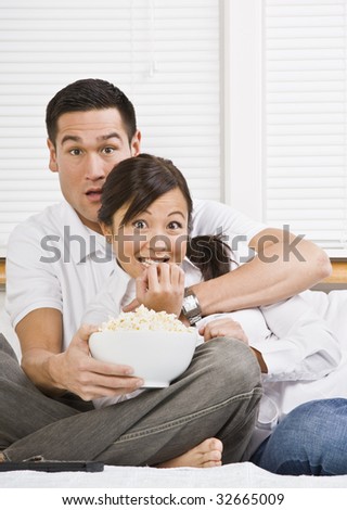 A young, attractive couple is sitting together in bed and watching TV.  They look shocked or scared, and are looking at the camera. Horizontally framed shot.