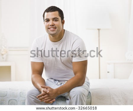A happy looking man seated on a bed in a white room.  He is smiling and looking at the camera.  Horizontally framed shot.