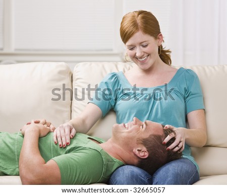 A cute young couple lounging on a couch together. The female is holding the male\'s head on her lap.  They are laughing and looking at one another. Square framed shot.
