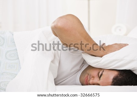 A tired looking man holding a pillow over his head and lying in bed.  Horizontally framed shot.