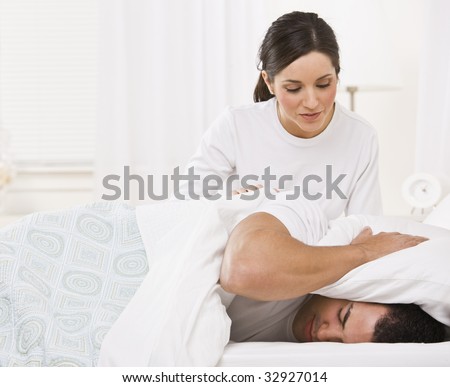 Attractive brunette woman trying to get her sleeping husband out of bed. Horizontally framed photo.