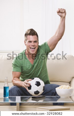 An attractive young man holding a soccer ball and cheering with his fist in the air.  He is looking directly at the camera. Vertically framed photo.