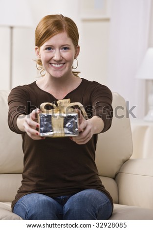 An attractive young female sitting on a couch and presenting a gift with outstretched arms.  She is looking directly at the camera and is smiling. Vertically framed photo.