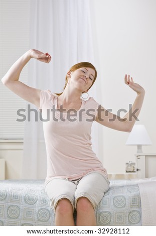 A young female stretching her arms.  She is sitting on a bed and is smiling.  Her eyes are closed.  Vertically framed photo.