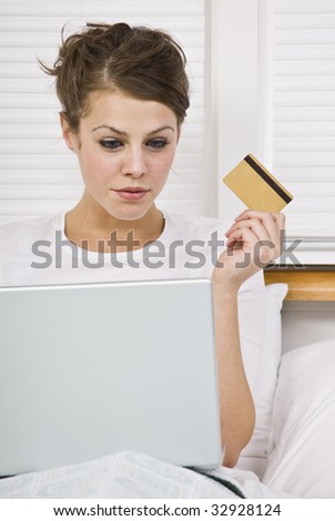 An attractive young woman holding a credit card and peering at a laptop screen.  She is sitting in bed and has a contemplative expression. Vertically framed shot.