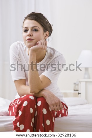 An attractive young woman sitting on the edge of a bed in her pajamas.  She is resting her chin on her hand and is smiling slightly toward the camera. Vertically framed photo.