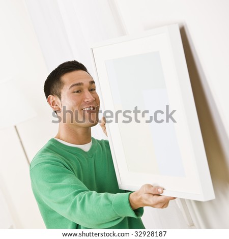 Asian male wearing a green sweater hanging art on wall. Square composition