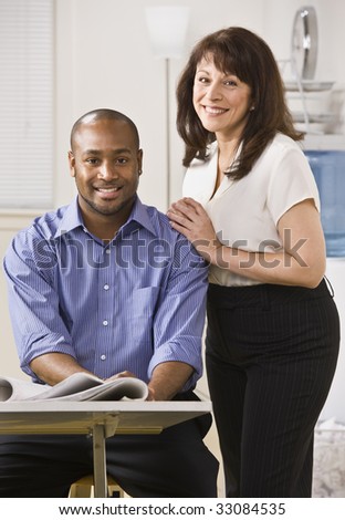 A young man is seated in an office and an older woman is standing next to him with her arms on his shoulder.  They are smiling at the camera.  Vertically framed shot.