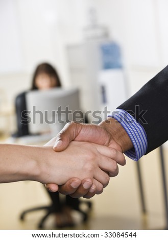 A businessman and woman are shaking hands in an office. There is another woman working on a computer in the background Vertically framed shot.