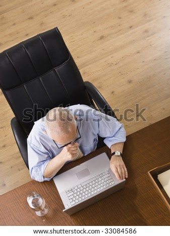 An elderly man is seated at a desk and is working on a laptop.  He is looking away from the camera.  Horizontally framed shot.