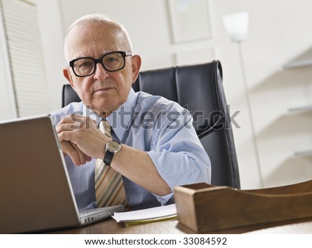 An elderly man is seated at a desk in front of a laptop and is looking at the camera.  Horizontally framed shot.
