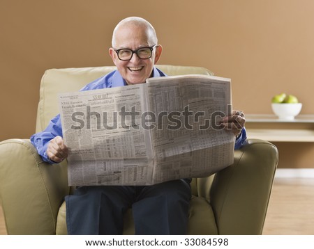 An elderly man is sitting in his living room reading a newspaper.  He is smiling at the camera.  Horizontally framed shot.