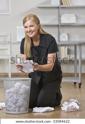 A businesswoman is kneeling at a trash can in an office and is putting all of the paper off the floor into it.  She is smiling at the camera.  Vertically framed shot.