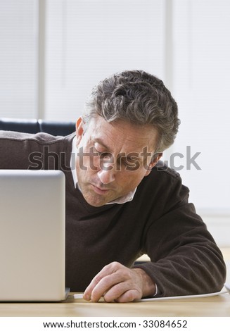 A businessman is seated at a desk in an office and is working on a laptop.  He is looking away from the camera.  Vertically framed shot.