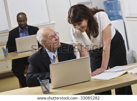 Attractive woman standing over older man helping him with laptop. African American male in back. Horizontal.