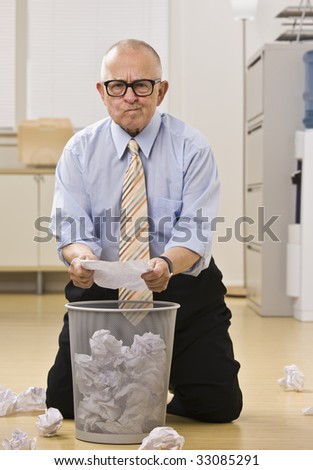 Attractive male senior kneeling on the floor, searching the garbage papers with questioning look, facing the camera. Verical.