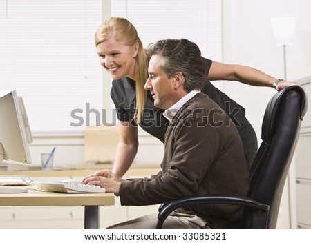 Attractive male and female working on computer. Female standing over male sitting at desk. Looking at computer. Horizontal