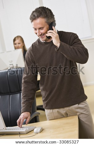 Attractive business man standing over keyboard while chatting on the phone. Woman peering in back. Looking at the computer screen. Vertical.