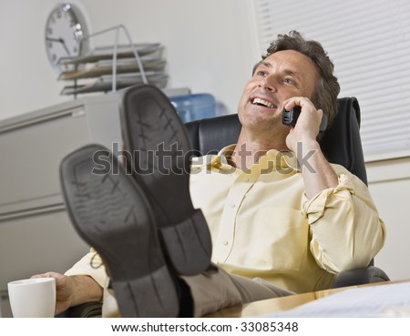 Attractive business man leaning back in chair, with legs on desk while chatting on the phone. Horizontal.