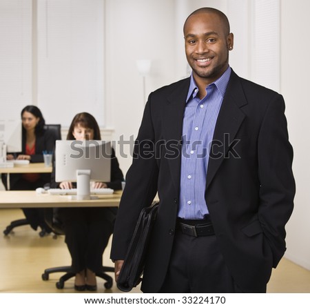 A young businessman is standing in an office with some other business people.  He is smiling at the camera.  Horizontally framed shot.