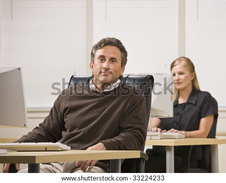 A businessman and woman are working in an office on computers.  The woman is looking at the computer screen and the man is looking at the camera.  Horizontally framed shot.