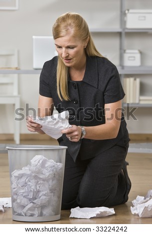 A businesswoman is on the floor in an office and is lookign through the trash.  She is looking away from the camera.  Vertically framed shot.