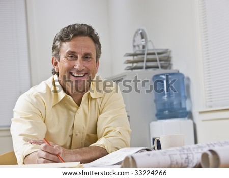 A businessman is seated at a desk in an office.  He is smiling at the camera.  Horizontally framed shot.