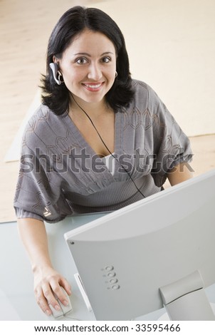 A young businesswoman is seated in front of a computer and is smiling at the camera.  Vertically framed shot.